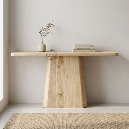 A light mango wood console table with a thick rectangular slab top and a tapered block base, styled with a small vase of dried stems and a stack of books against a neutral wall.