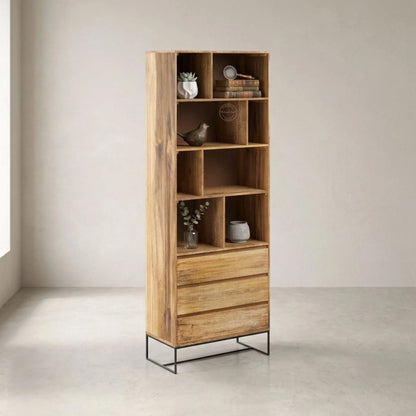 A tall, narrow mango wood bookcase with an open grid upper section, three drawers at the bottom, and a black metal wire frame base, styled with home decor items in a minimalist room.