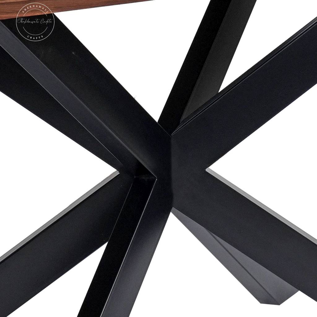 Close-up view of a black metal table base with angular intersecting legs on a white background, showing part of a wood tabletop edge at the top.
