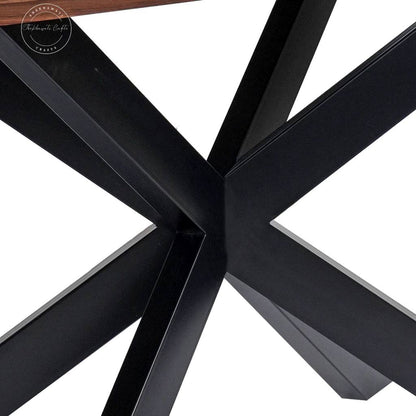 Close-up view of a black metal table base with angular intersecting legs on a white background, showing part of a wood tabletop edge at the top.