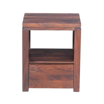 A wooden bedside table made of sheesham wood with a honey finish, featuring a drawer and an open shelf.