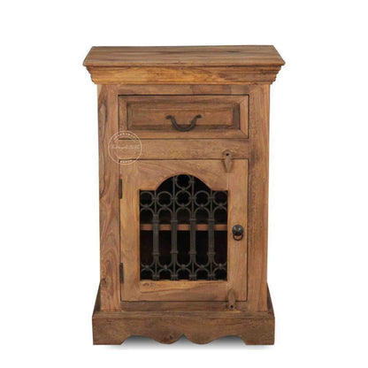 A wooden bedside table with a drawer, a shelf, natural finish and an iron jali door in a white background.