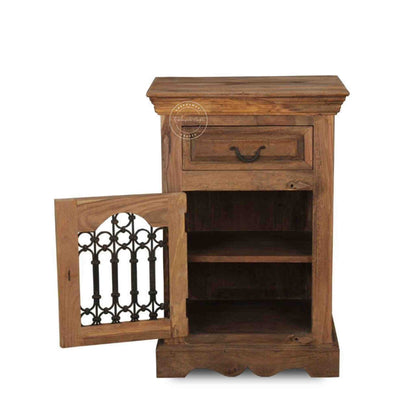 A wooden bedside table with a drawer, a shelf, natural finish and an iron jali door in a white background.
