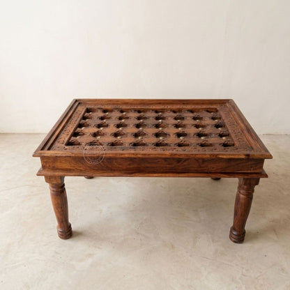A rectangular dark brown wooden table with carved details and a grid of circular cutouts on the top, supported by four turned legs.