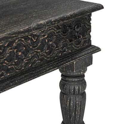 Close-up of a dark distressed wooden table corner showing carved floral details on the apron and a turned pedestal leg, on a white background.