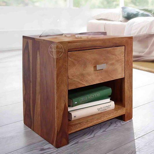 A wooden Zoey Bedside table with one drawer and an open shelf, placed on a floor in a room with books on the shelf.
